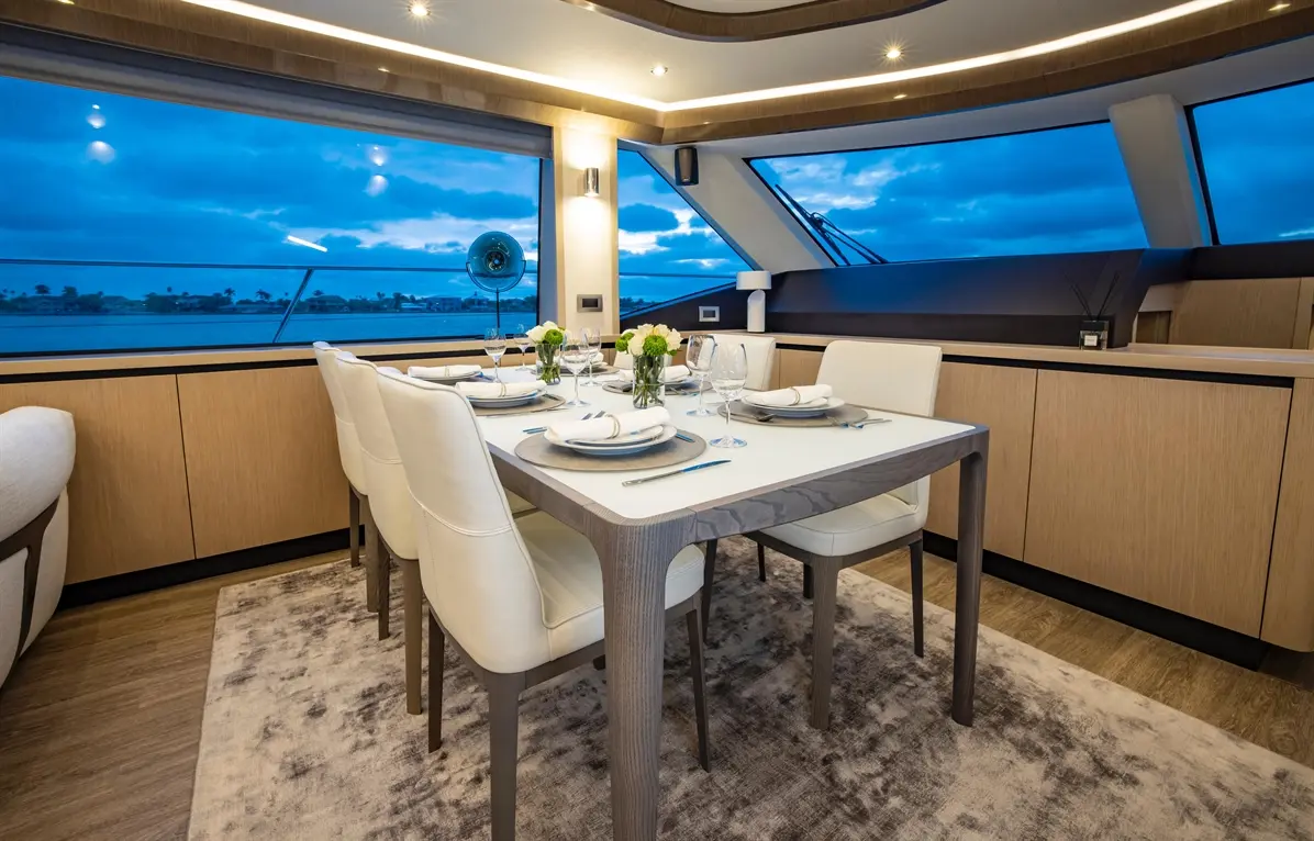 Modern dining area with a table set for six, large windows showing a blue sky and water view, adorned with floral centrepieces.